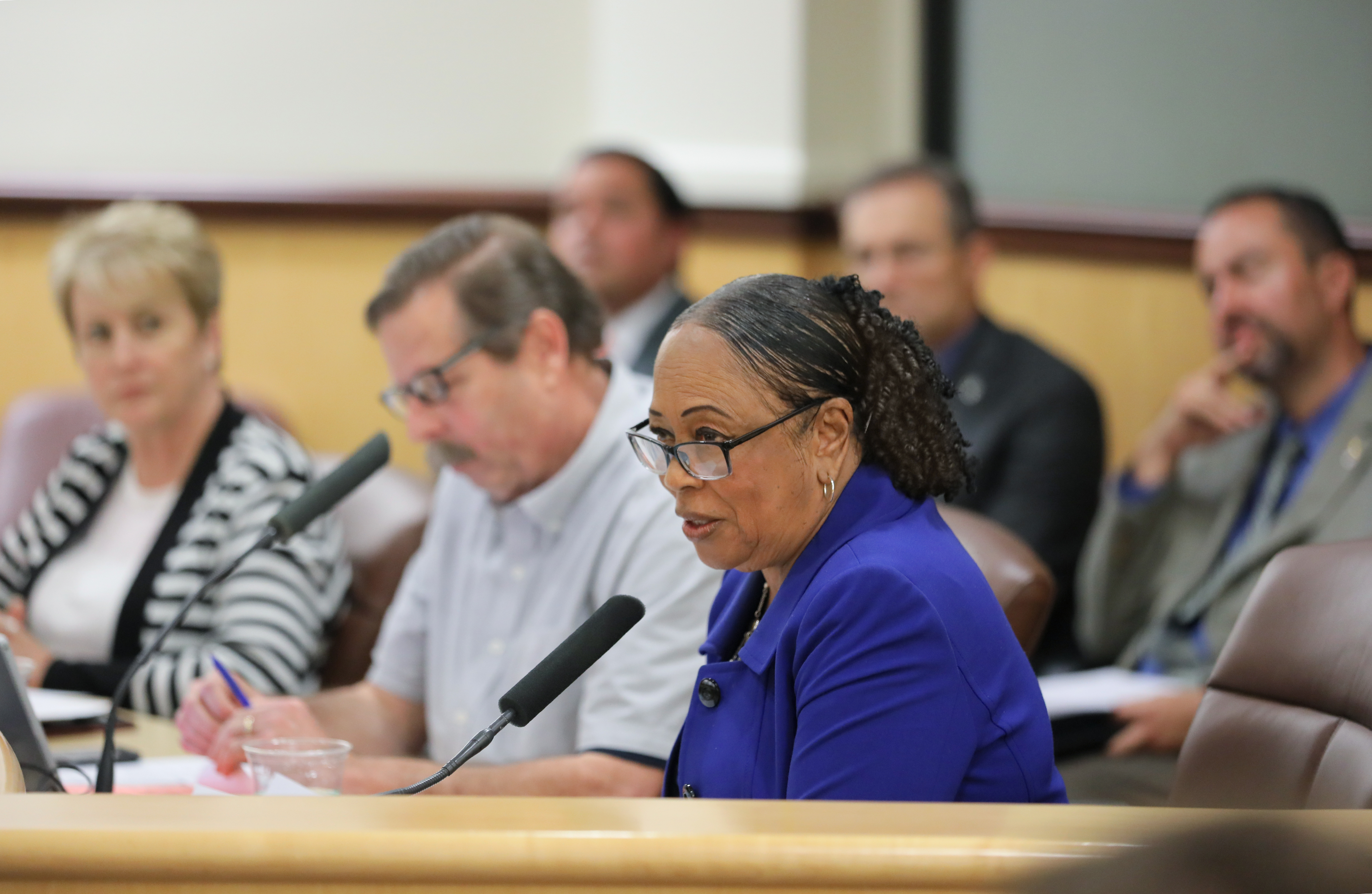 Six people in two rows of three. The front row is sitting in front of microphones and is talking to the rest of the room off frame.