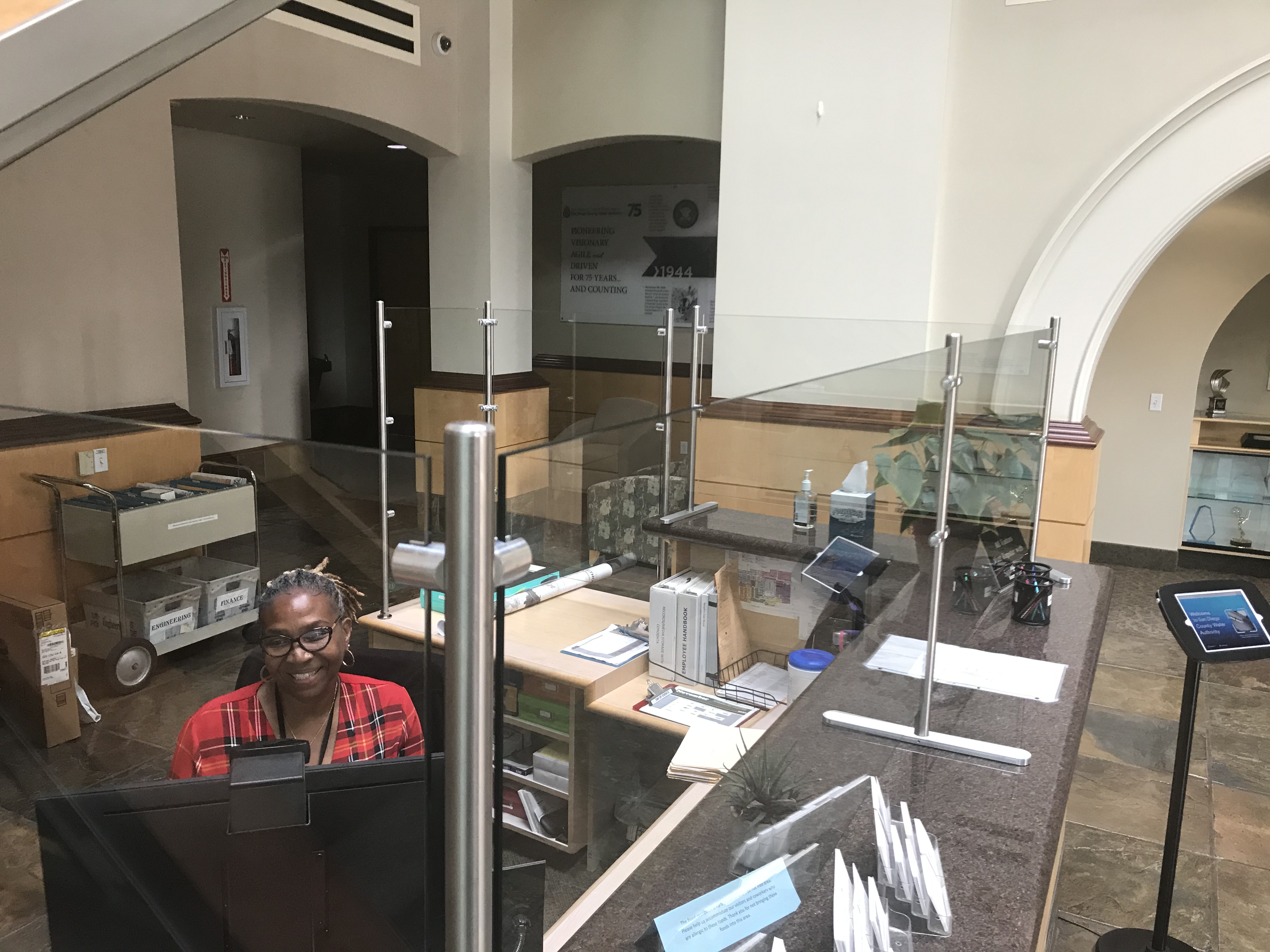 Building lobby with reception desk, check-in and check-out screen and transparent shields at the counter.
