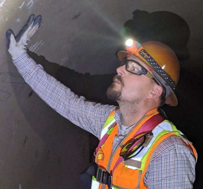 A man wearing a hardhat and construction vest stands inside a large-diameter water pipe.