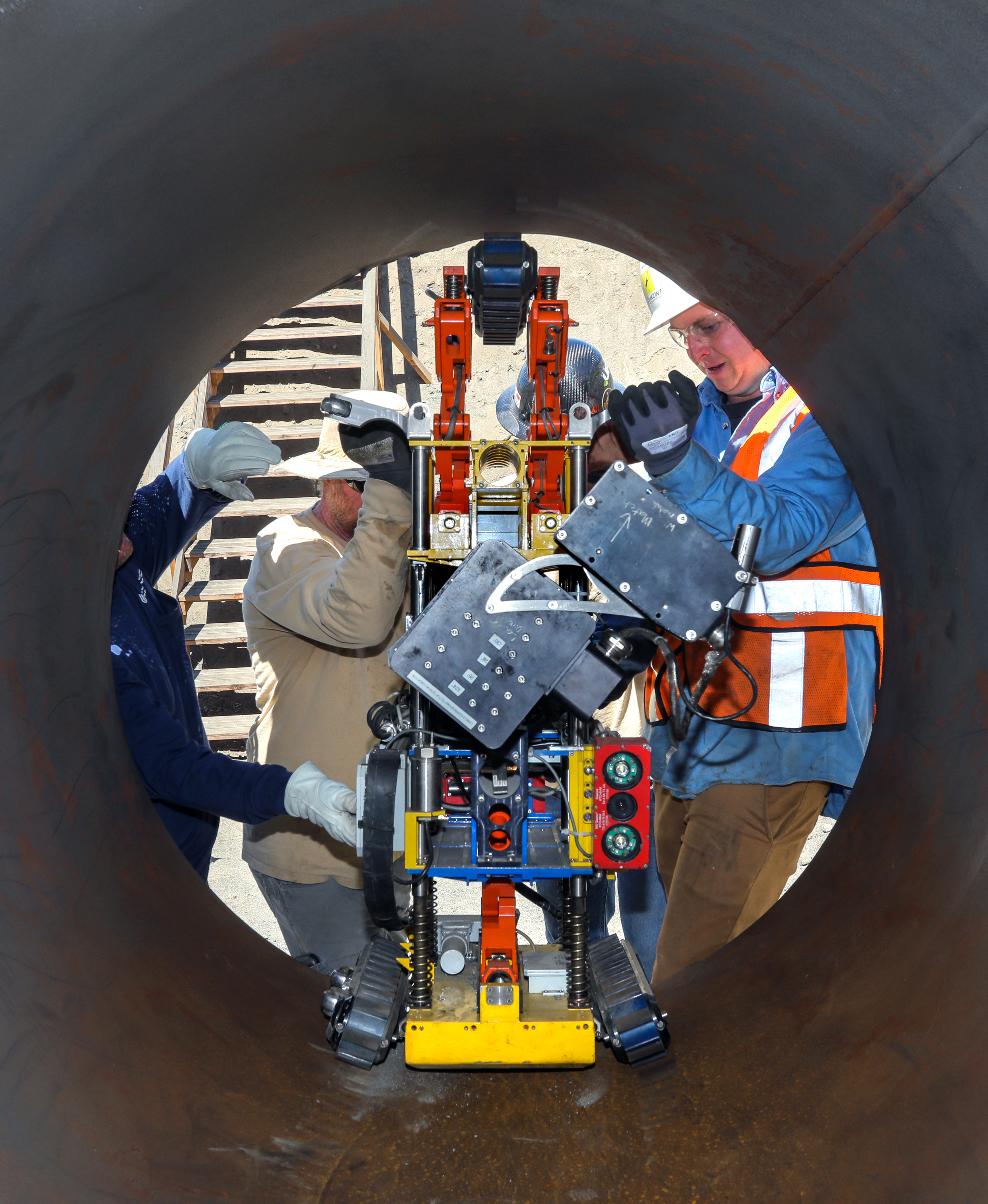 An employee in a hard hat and construction vest holds a robot inside a water pipeline that has had a section removed for rehabilitation.